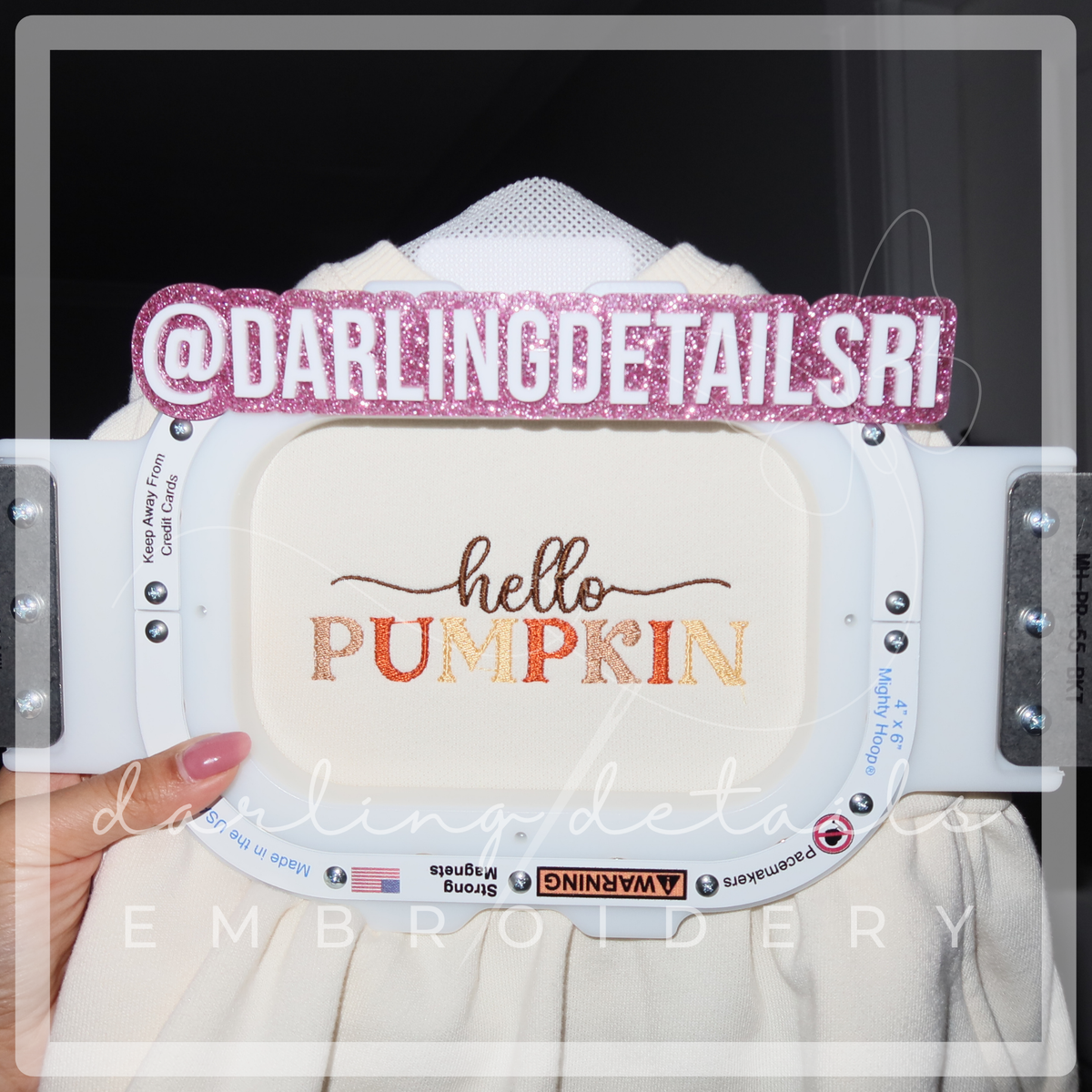 A close-up of an embroidery hoop holding a cream fabric with the phrase 'hello PUMPKIN' embroidered in brown and orange. A pink glittery '@DARLINGDETAILSRI' sticker is placed across the hoop, and a hand with pink nail polish holds the left side.