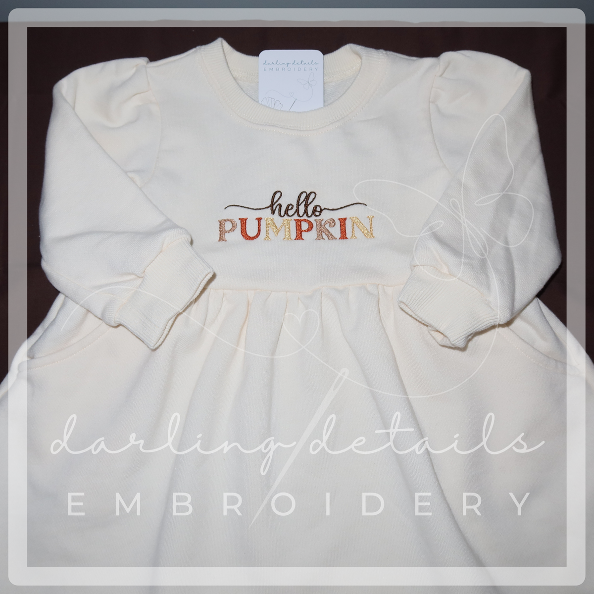 An off-white long-sleeve dress with 'hello PUMPKIN' embroidered on the chest, laid flat on a dark background with a watermark overlay.