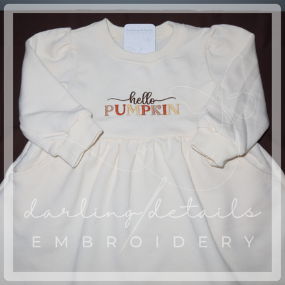 An off-white long-sleeve dress with 'hello PUMPKIN' embroidered on the chest, laid flat on a dark background with a watermark overlay.