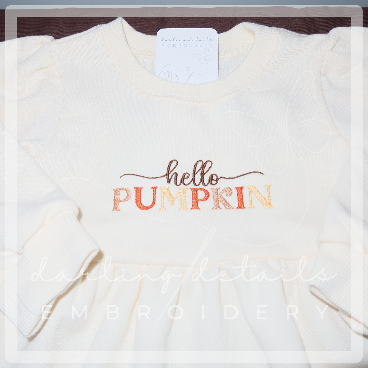 An off-white long-sleeve dress or featuring 'hello PUMPKIN' embroidered on the chest. 'hello' is in brown cursive, and 'PUMPKIN' is in orange and yellow textured letters.