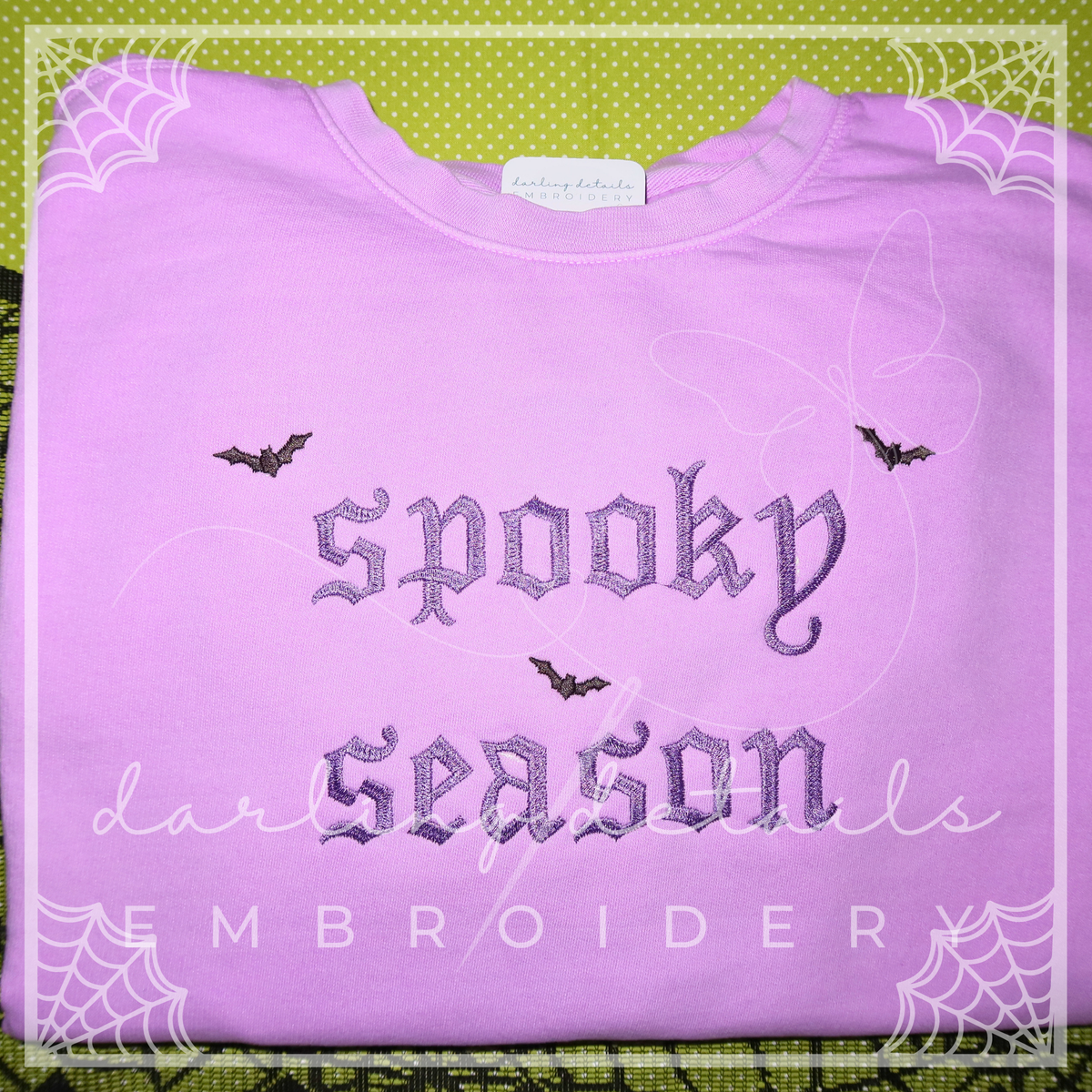 A folded light purple sweatshirt with gothic-style embroidered text reading 'spooky season' and small bat motifs, displayed on a green patterned background with decorative spiderweb borders.
