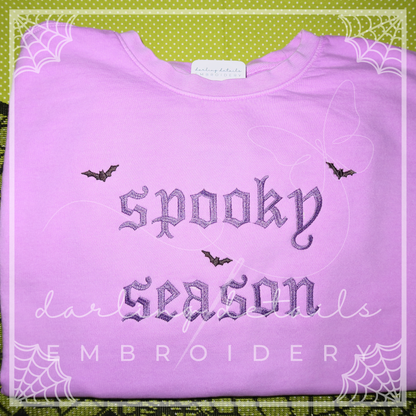 A folded light purple sweatshirt with gothic-style embroidered text reading 'spooky season' and small bat motifs, displayed on a green patterned background with decorative spiderweb borders.