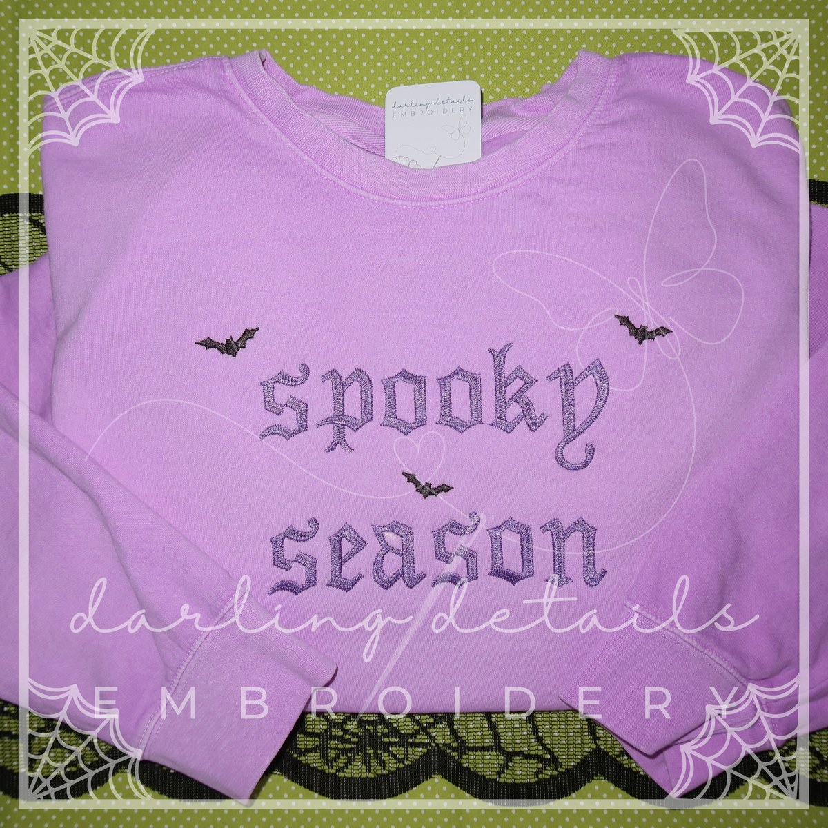 A folded lavender crewneck sweatshirt featuring 'spooky season' embroidered in gothic-style lettering with small bat motifs. The sweatshirt is displayed on a green patterned background with decorative spiderweb borders and a watermark overlay.