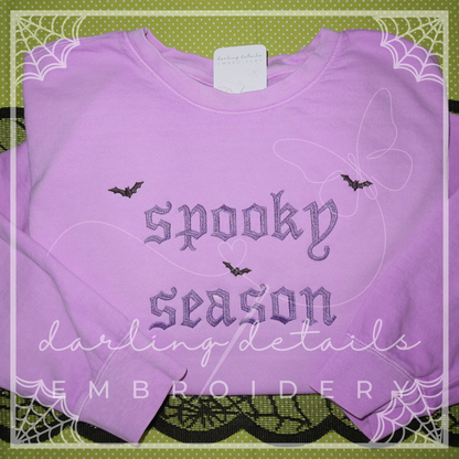 A folded lavender crewneck sweatshirt featuring 'spooky season' embroidered in gothic-style lettering with small bat motifs. The sweatshirt is displayed on a green patterned background with decorative spiderweb borders and a watermark overlay.