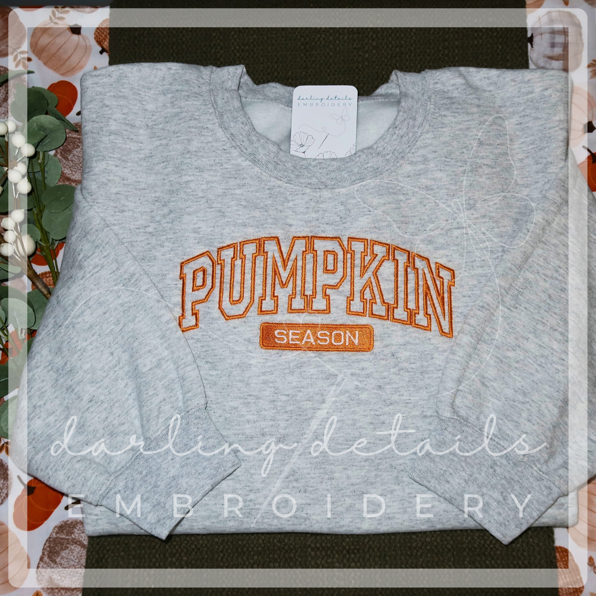 A folded heather gray crewneck sweatshirt with 'PUMPKIN SEASON' embroidered in orange and brown on the front, displayed flat on a dark surface with a patterned fabric and decorative leaves around it.