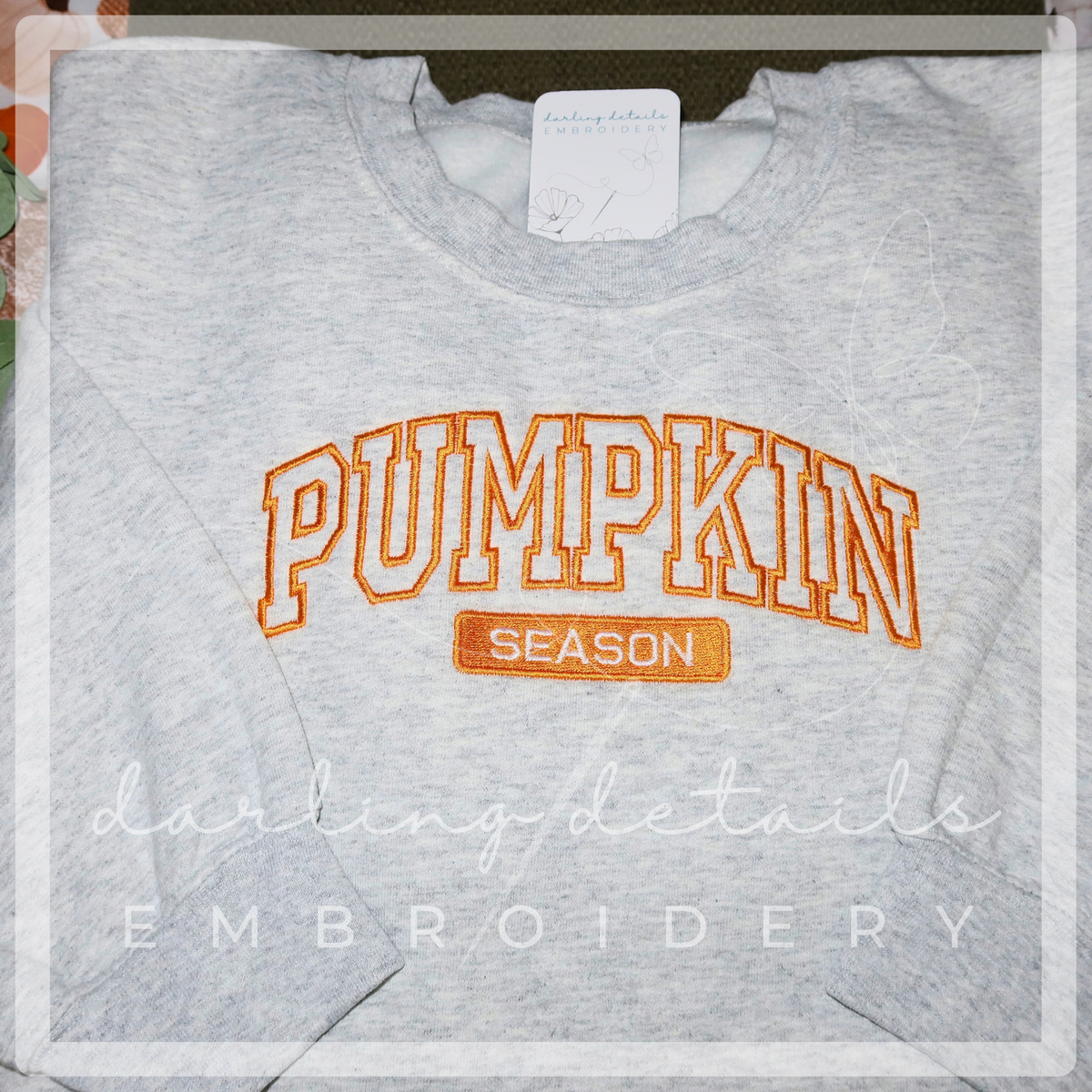 A light heather gray crewneck sweatshirt featuring 'PUMPKIN SEASON' embroidered in orange on the front, with a white tag visible inside the collar and a decorative watermark overlay.