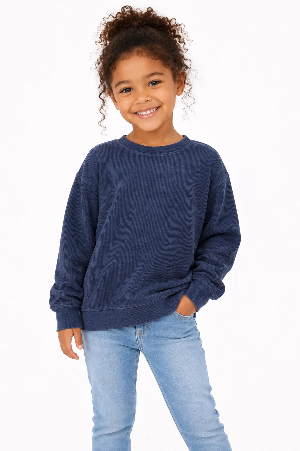 A young girl with curly brown hair smiling, wearing a dark navy blue crewneck sweatshirt and light blue jeans, standing against a plain white background.