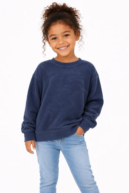 A young girl with curly brown hair smiling, wearing a dark navy blue crewneck sweatshirt and light blue jeans, standing against a plain white background.