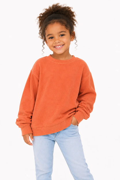 A young girl with curly brown hair wearing a washed orange crewneck sweatshirt and light blue jeans, standing against a plain white background.