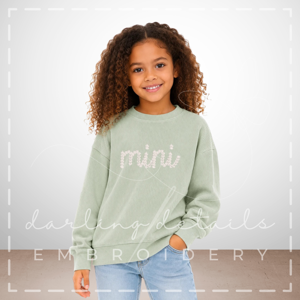 A young girl with curly brown hair wearing a sage green crewneck sweatshirt with 'mini' embroidered in off-white across the chest, paired with light blue jeans, standing against a light gray wall.